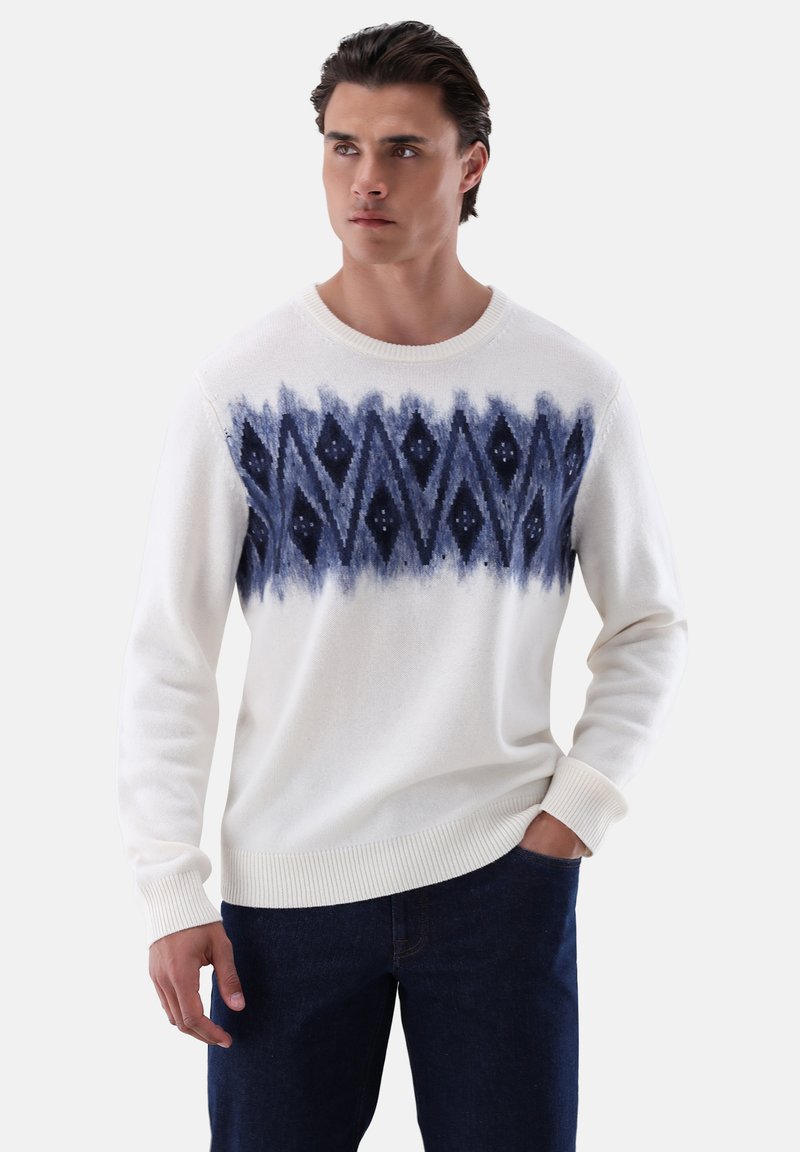 Weißer Strickpullover mit einem navyblauen Mustermuster, das Rauten und geometrische Formen zeigt. Mit Rippenbündchen und Saum, getragen mit dunklen Jeans.
