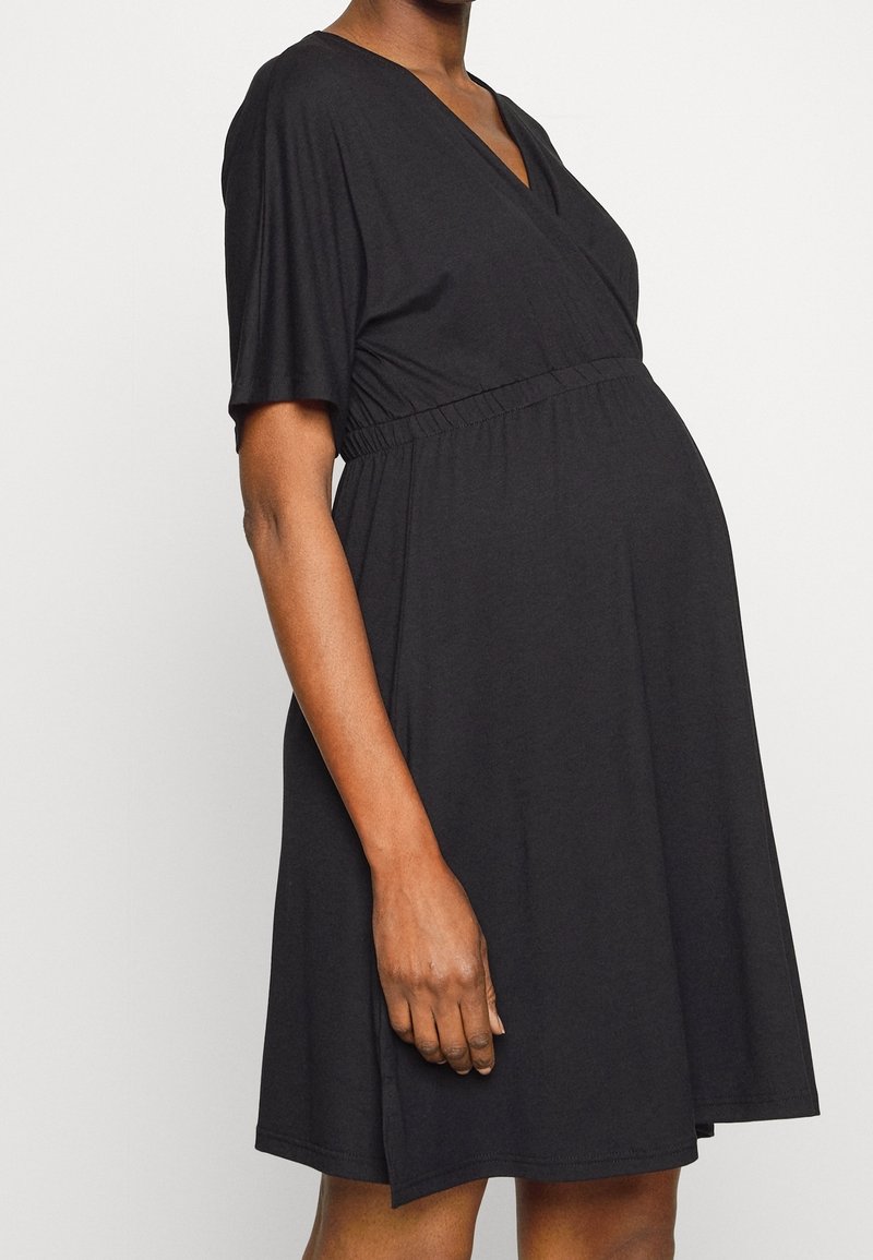 Czarna sukienka ciążowa wykonana z miękkiego materiału; ma dekolt w serek, krótkie rękawy i wysoki stan. Spódnica ma luźny krój.