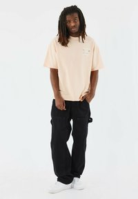 Licht perzik oversized t-shirt en zwarte cargobroek. Het t-shirt heeft subtiele grafische details. Het model staat met de handen in de zakken.