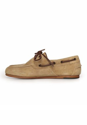 Beige suède mocassins met donkerbruine leren veterdetails en platte bruine zool, zijaanzicht op een witte achtergrond.