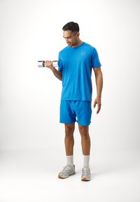 Helles blaues Sport-T-Shirt und Shorts, der Stoff scheint leicht zu sein; hält eine Metall-Hantel, trägt graue Sneakers und weiße Socken.