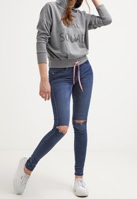 Sudadera gris con la impresión "SQUAD", combinada con unos jeans ajustados de color azul oscuro con rasgaduras en las rodillas, y zapatillas blancas con superficies texturizadas.