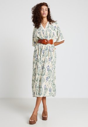 Donna con capelli ricci indossa un vestito midi a V con fantasia, stretto in vita da una larga cintura marrone e sandali con zeppa marroni, in piedi contro un muro semplice.