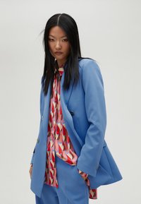 Blazer doppiopetto blu chiaro con bottoni argentati, indossato sopra una blusa a fantasia rossa e rosa con forme geometriche.