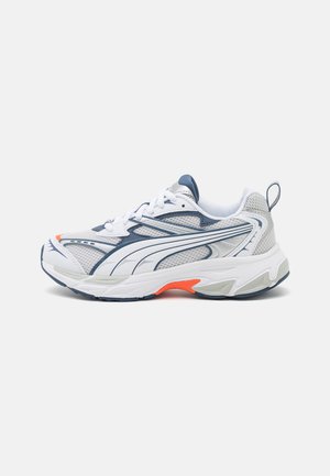 Scarpa atletica bianca con accenti blu navy e arancioni, tomaia in rete, sovrapposizioni testurizzate, suola in gomma e lacci. Presenta un linguetta posteriore.