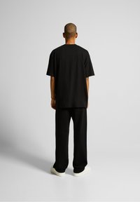 Camiseta negra oversize de manga corta, combinada con pantalones negros de corte holgado y zapatillas blancas, vista desde la parte de atrás. Diseño sencillo.