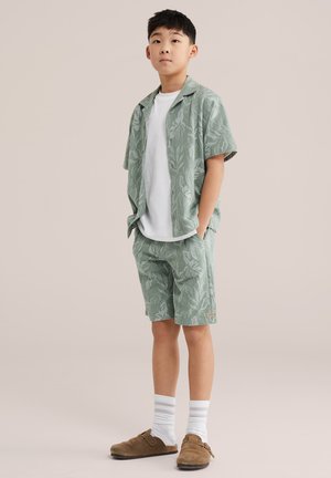 Boy standing with hands in pockets, wearing green leaf-patterned short-sleeve shirt and matching shorts, white T-shirt, white socks, and brown slip-on shoes.