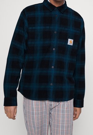 Man draagt een blauw-zwart geruit overhemd met logo op de borstzak en geruite broek met witte, zwarte en rode strepen.