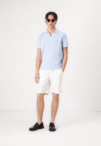 Tricou polo cu mânecă scurtă, de culoare bleu deschis, cu tricot texturat, pantaloni scurți albi și pantofi negri fără șireturi. Ochelarii de soare completează ținuta.