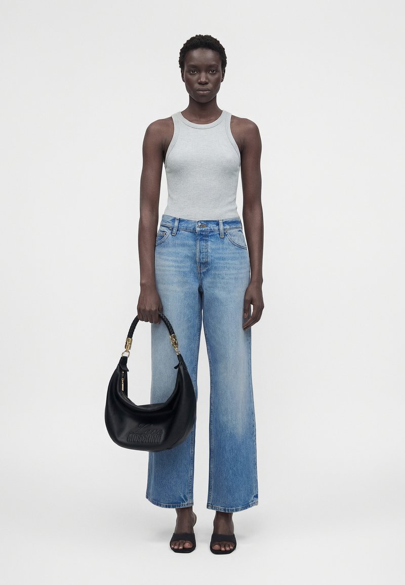 Γυναίκα στέκεται φορώντας ανοιχτό γκρι αμάνικη μπλούζα, μπλε τζιν παντελόνι με ευρύ μπατζάκι, μαύρα πέδιλα με ανοιχτά δάχτυλα, κρατώντας μια μαύρη τσάντα με πλεκτή λαβή.