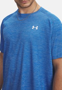 Camiseta deportiva azul con un diseño texturizado, de mangas cortas y con un logotipo blanco de Under Armour en el pecho. Tejido suave con una sensación ligera.