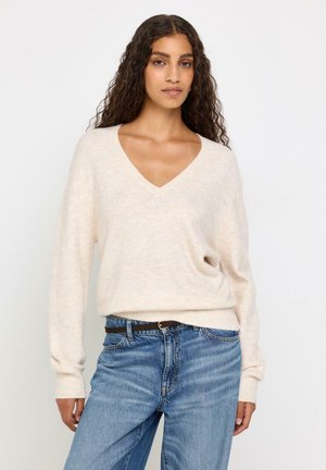 Donna con lunghi capelli ricci che indossa un maglione crema a scollo a V infilato in jeans blu con cintura nera su uno sfondo bianco semplice.