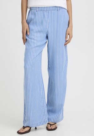 Kobieta ubrana w jasnoniebieskie spodnie o szerokich nogawkach z białymi pionowymi paskami oraz brązowe sandały na obcasie, stojąca na jednolitym tle.