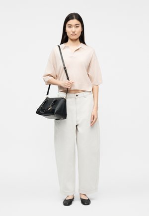 Jonge vrouw met lang donker haar draagt een loszittend lichtroze overhemd, brede lichtbeige broek, zwarte platte schoenen en draagt een zwarte handtas over haar schouder.