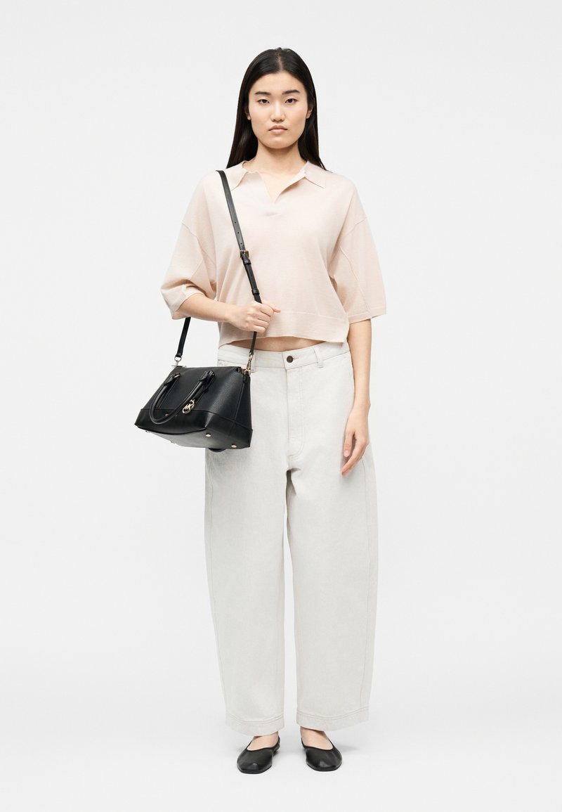 Νεαρή γυναίκα με μακριά σκούρα μαλλιά φοράει ένα φαρδύ ανοιχτό ροζ πουκάμισο, φαρδιά ανοιχτό μπεζ παντελόνια, μαύρα μπαλαρίνια και κρατά μια μαύρη τσάντα στον ώμο της.