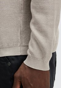 Nahaufnahme einer Person, die einen strukturierten beigefarbenen Pullover mit gerippten Bündchen und dunklen Hosen trägt, wobei das Strickmuster und die Passform des Pullovers zu sehen sind.