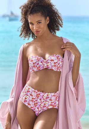 Frau mit lockigem Haar, die einen pinkfarbenen, geblümten Trägerlosen Bikini und ein hellrosa Überwurf trägt, steht am türkisfarbenen Ozean.