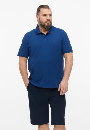 Mężczyzna z brodą ubrany w niebieską koszulkę polo z krótkim rękawem i ciemne szorty, stoi z rękami w kieszeniach na jednolitym tle.