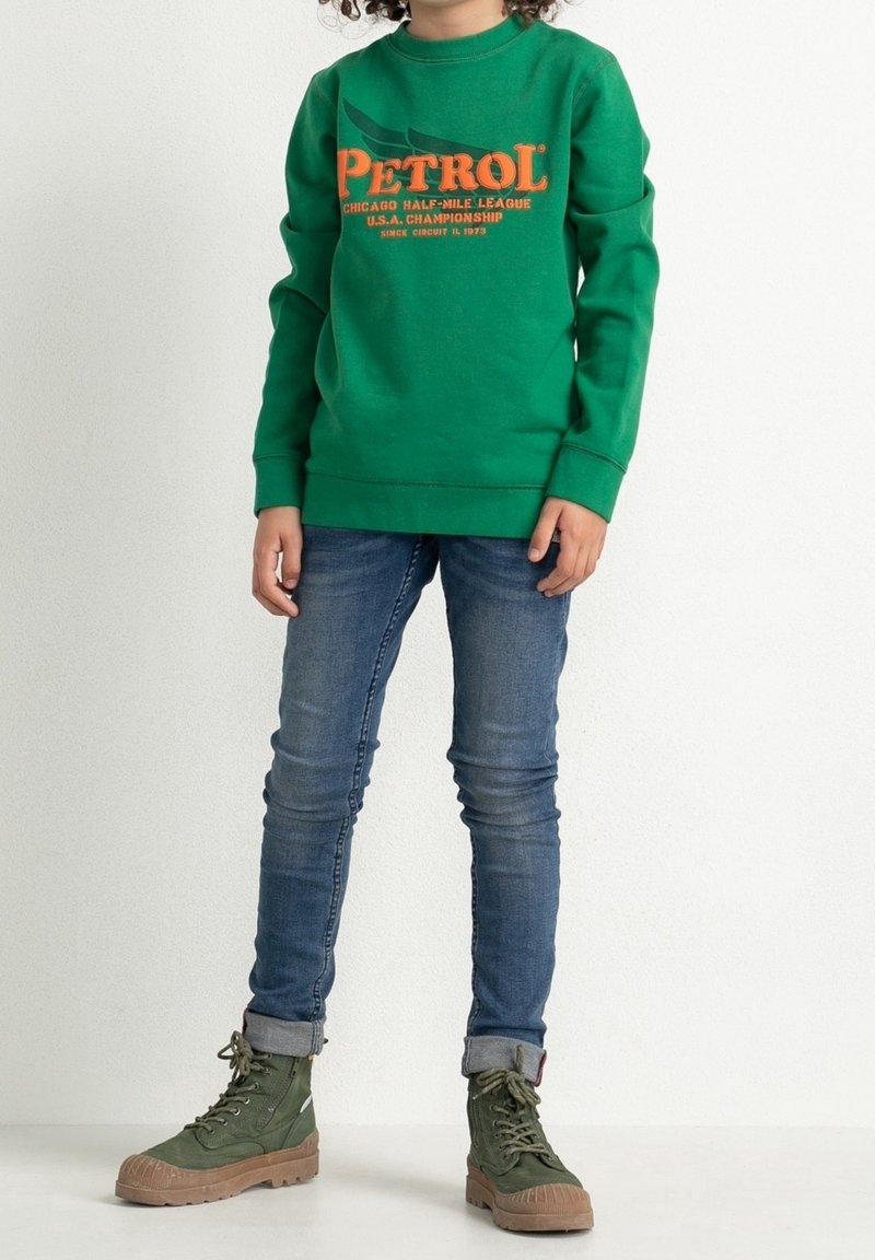 Barn i grön sweatshirt med orange text, blå uppvikt jeans och gröna snörstövlar, står framför en vit bakgrund.