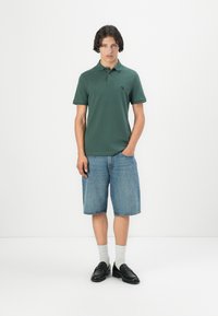 Chemise polo verte à manches courtes avec un logo noir, associée à un short en denim. Portée avec des chaussettes gris clair et des mocassins noirs.