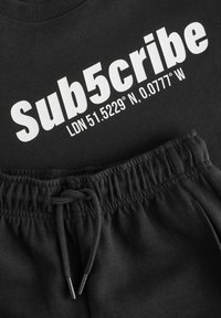 Sort sweatshirt med hvid tekst "Sub5cribe LDN 51.5229° N, 0.0777° W," parret med sorte sweatpants med snøre, der har en blød tekstur.