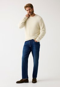 Suéter texturizado de color crema combinado con jeans azul oscuro y mocasines marrones; pose de pie sobre un fondo neutro.