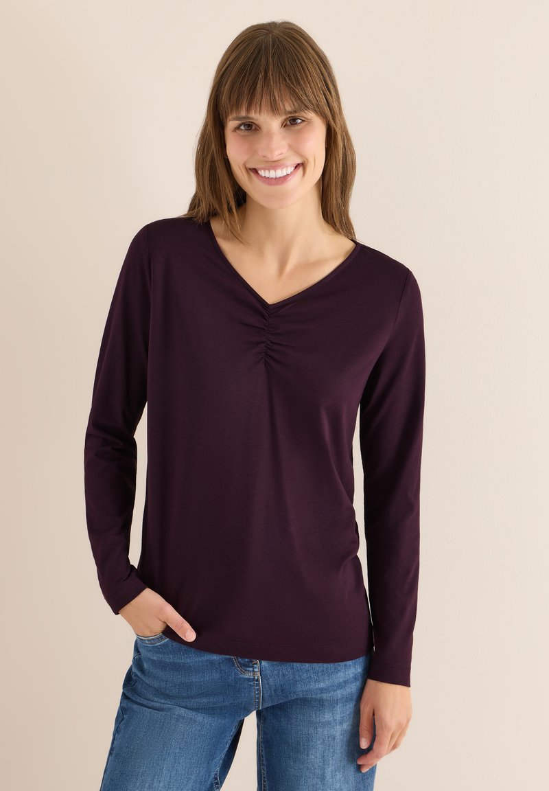 Long-sleeve, deep purple top with a V-neck and gathered detail at the center, paired with blue denim jeans. Soft fabric texture.