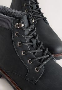 Cizme negre din piele cu guler tricotat gri texturat. Caracteristicile includ ochiuri metalice, șireturi negre și o talpă maro cusută.