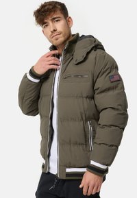 Olive green padded jacket with a hood, front zipper closure, two zippered pockets, and striped cuffs. Features an American flag patch on the sleeve.