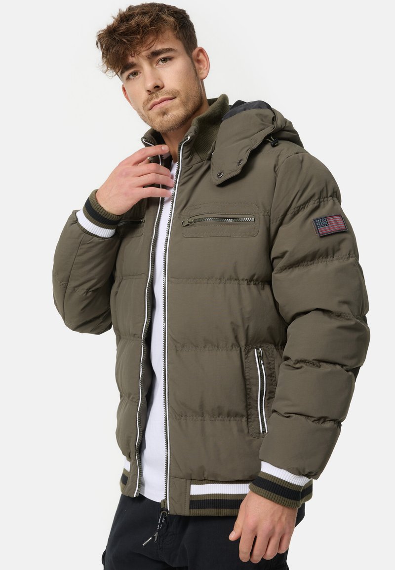 Olive green padded jacket with a hood, front zipper closure, two zippered pockets, and striped cuffs. Features an American flag patch on the sleeve.