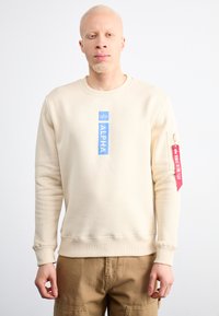 Bézs pulóver kerek nyakkal, elöl kék "ALPHA" felirattal és piros címkével a bal ujján. Puha tapintású, laza szabású.