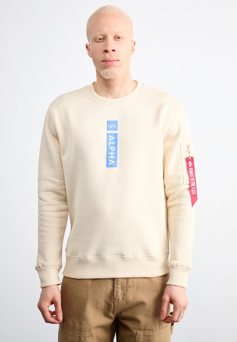 Bézs pulóver kerek nyakkal, elöl kék "ALPHA" felirattal és piros címkével a bal ujján. Puha tapintású, laza szabású.