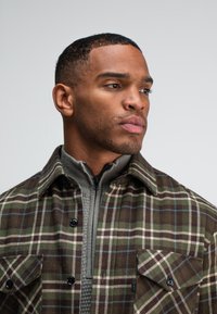 Geruite shirt in aardetinten, met groene, bruine en witte patronen, zwarte knopen en een hoge kraag grijze ritslaag eronder.