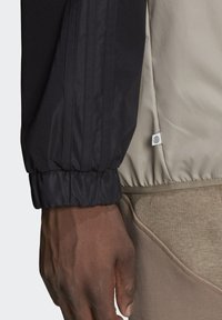 Närbild av en persons hand som bär en svart jacka med elastiskt ärm, en beige jacka och bruna byxor med synlig söm vid sidofickan.