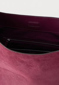 Interiér ružovej semišovej tašky na rameno, ktorý obsahuje vrecko so zipsom a hladké kožené detaily. Zaoblené hrany a jemná textúra zlepšujú dizajn.