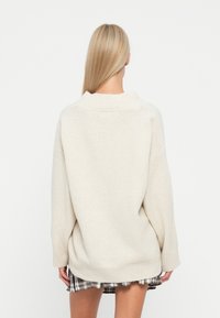 Maglione in maglia oversize di un beige chiaro con colletto alto e orlo a coste. La vista posteriore mostra una vestibilità morbida e un tessuto testurizzato. Abbinato a una gonna a quadri.