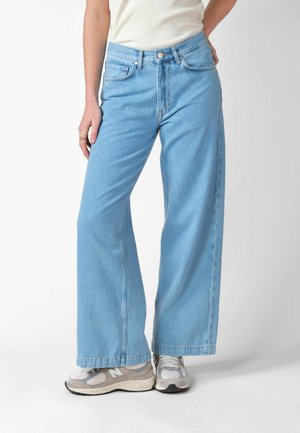 Osoba ubrana w jasnoniebieskie szerokie spodnie dżinsowe, biały top na ramiączkach i szare buty sportowe New Balance, stojąca na tle jednolitego białego tła.