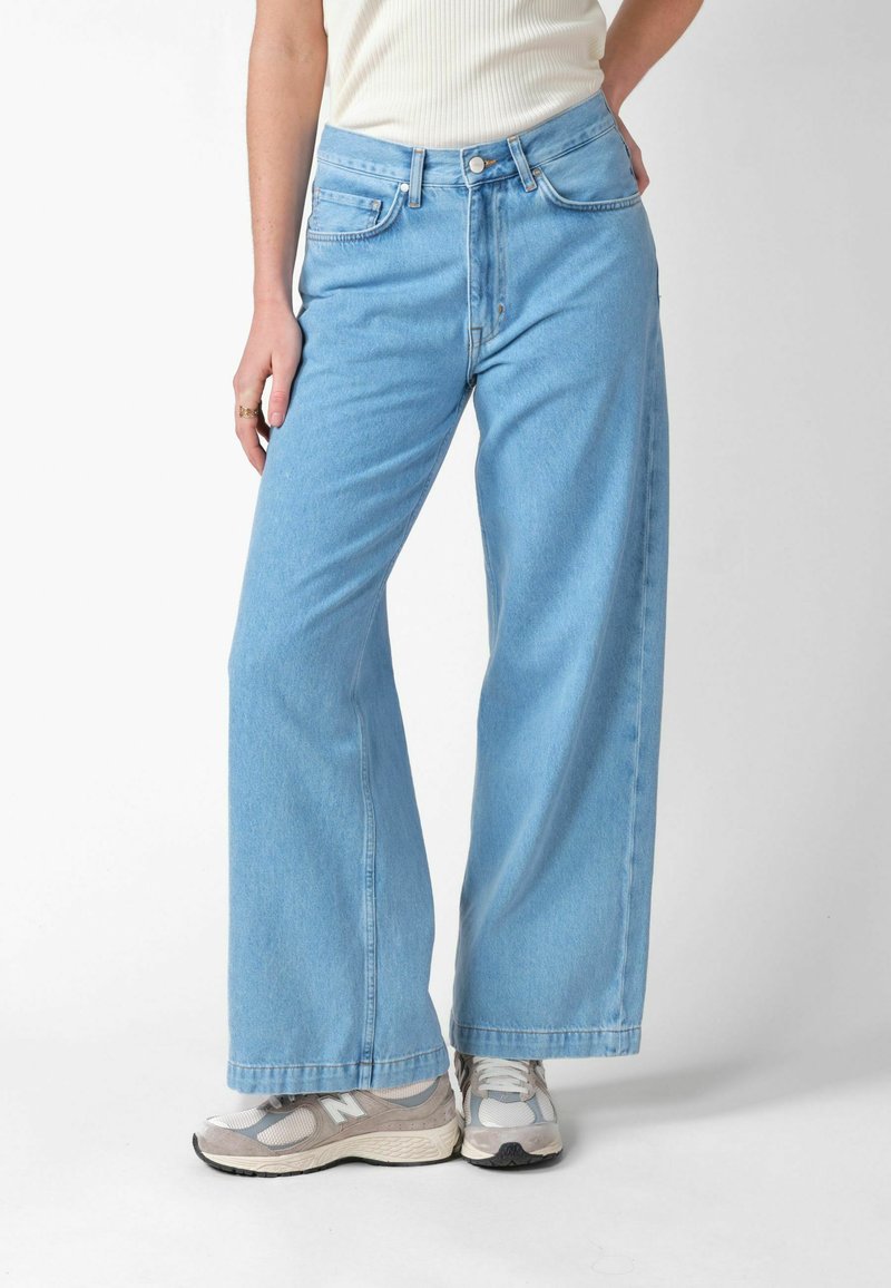 Personne portant un jean large bleu clair, un débardeur blanc et des baskets New Balance grises, debout devant un fond blanc uni.