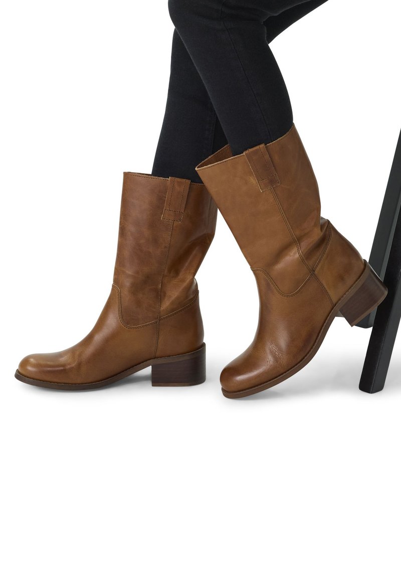 Bottines en cuir marron avec un petit talon carré, texture lisse et un design légèrement tombant. Couture visible et accents latéraux.
