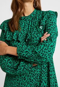 Groene jurk met zwarte luipaardvlekken, met geplooide halslijn en mouwen, lange mouwen en knoopmanchetten. Zachte, gestructureerde stof.