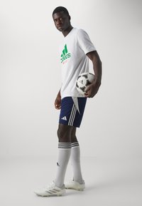 Bijela Adidas majica s zelenim logotipom, tamnoplave kratke hlače, bijele nogometne čarape s crnim prugama i bijeli nogometni kopački, držeći crno-bijelu nogometnu loptu.
