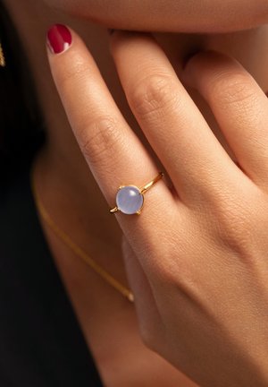 Goldring mit einem runden, hellblauen Edelstein vor einem strukturierten Hauthintergrund, während die Finger sanft das Kinn berühren.