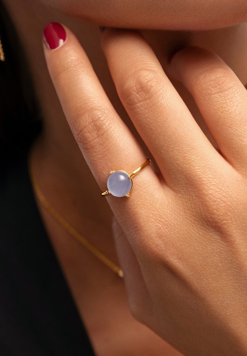 Goldring mit einem runden, hellblauen Edelstein vor einem strukturierten Hauthintergrund, während die Finger sanft das Kinn berühren.