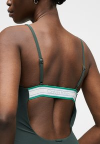 Bakside av kvinne som har på seg mørkegrønn topp med justerbare stropper, åpen rygg og hvitt Calvin Klein-merket bånd over øvre rygg.