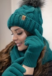 Gorro de punto verde aguamarina con un pompón de piel, guantes a juego de color verde aguamarina y una bufanda texturizada. El material parece cálido y acogedor con patrones acanalados.
