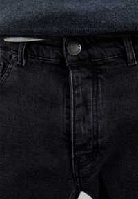Primer plano de unos vaqueros de denim negros con un botón metálico grabado "Scalpers" y un suéter de punto gris oscuro metido por dentro.
