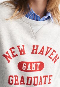 Sudadera gris con texto rojo que dice "NEW HAVEN GANT GRADUATE." Cuenta con un cuello redondo y tela ligeramente texturizada.