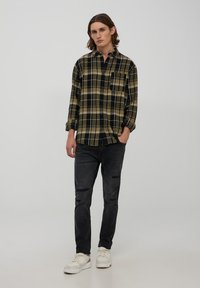 Camicia a quadri verde, nera e gialla con taschino sul petto, abbinata a jeans scuri strappati e sneakers chiare.