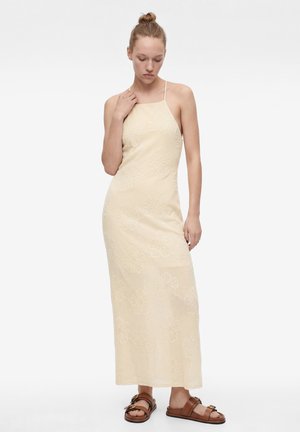 Vrouw draagt een mouwloze, crèmekleurige maxi-jurk met bloemenpatroon en bruine platte sandalen, staand tegen een effen witte achtergrond.