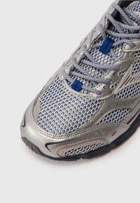 Sapatilha desportiva prata e azul com parte superior em malha, detalhes texturizados e sola de borracha resistente, com um intrincado design de atacadores.
